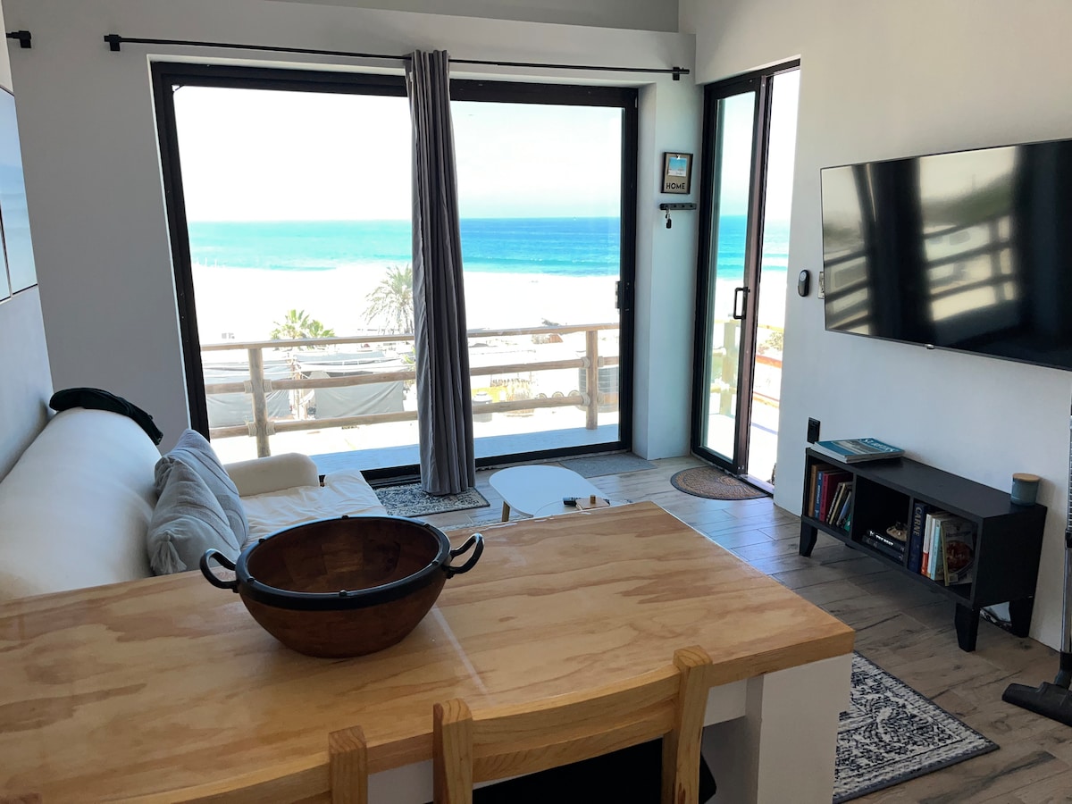 A spacious living area is shown with large glass doors offering a view of the ocean. Natural light fills the room, and a wooden dining table is set with a bowl in the center. A comfortable couch and a flat-screen TV complete the inviting space.