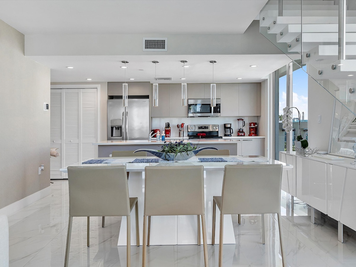 A modern dining area features a white table surrounded by six pale chairs. Above, pendant lights provide illumination. The fully equipped kitchen includes stainless steel appliances and a sleek countertop, with large windows offering natural light and views of the surrounding area.