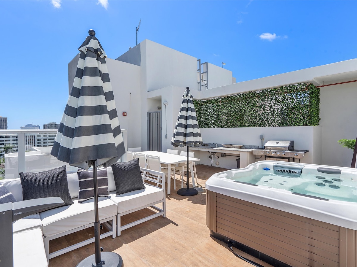 The rooftop terrace features comfortable lounge seating under large striped umbrellas. A private jacuzzi is positioned beside a grilling area, surrounded by greenery. Bright blue skies provide a serene backdrop, enhancing the outdoor space's inviting atmosphere.