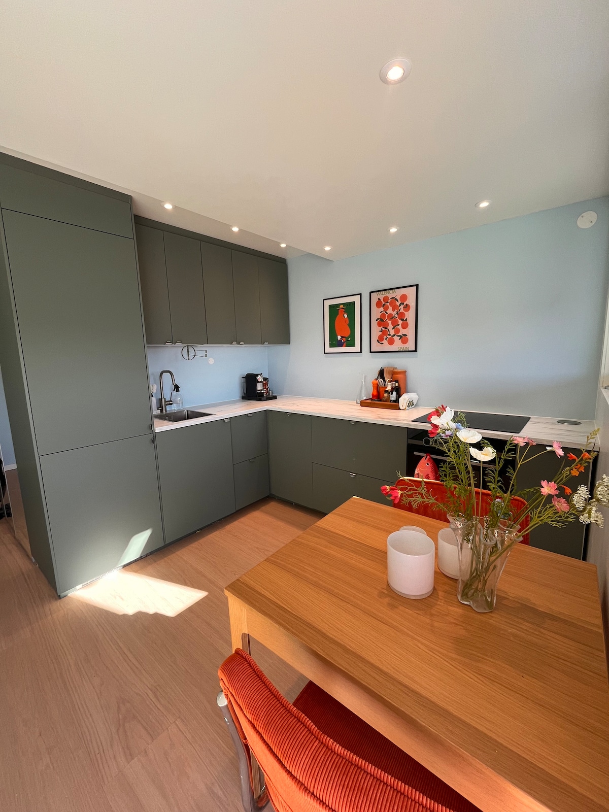 A modern kitchen features sleek cabinetry and contemporary appliances, including a coffee machine. A small dining table is set with a vase of flowers, adding a touch of color to the space. Natural light brightens the area from a nearby window.