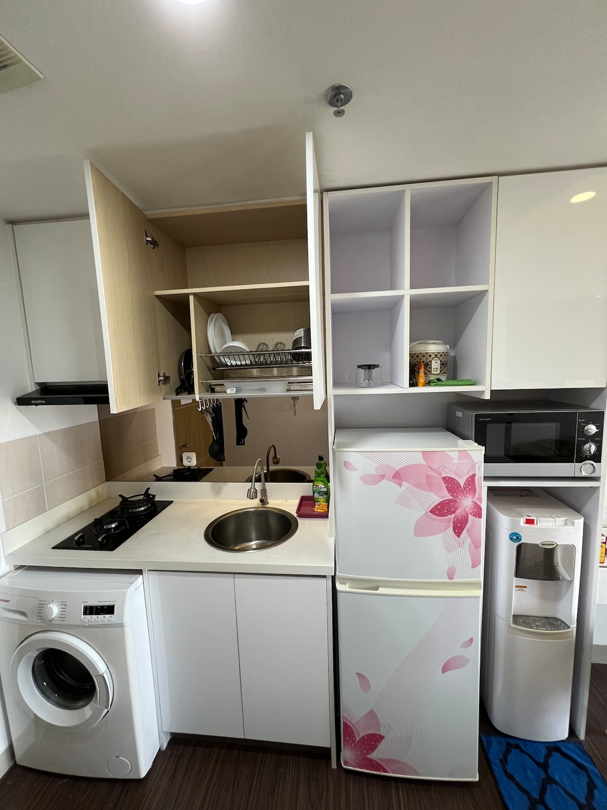 A compact kitchen area features a sink, stovetop, and under-counter appliances including a washing machine and refrigerator. Shelves above hold dishware, while a microwave is positioned on the countertop. The space is designed with a clean, modern aesthetic highlighted by a floral pattern on the fridge.