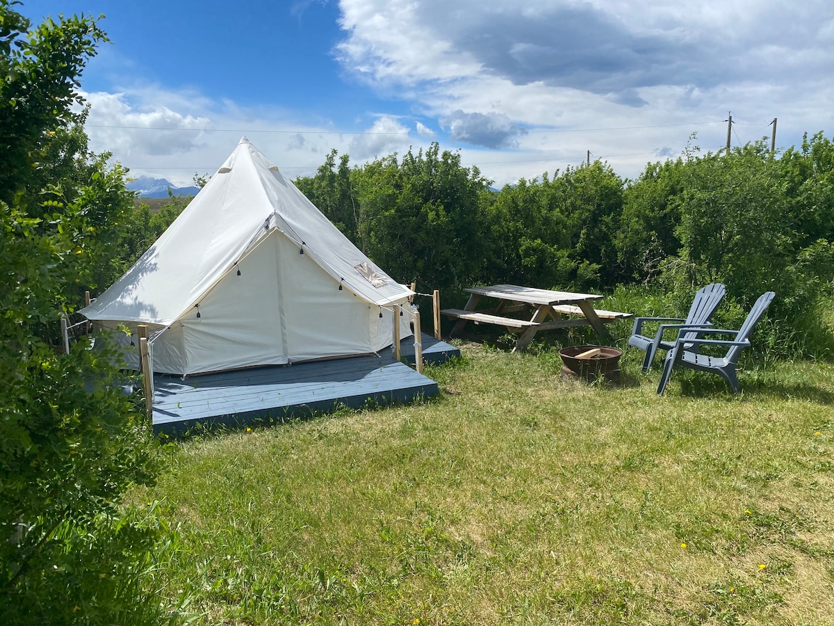 A spacious canvas tent is set on a wooden platform, surrounded by lush greenery. A picnic table and two adirondack chairs are positioned nearby, along with a small fire pit. The sky is partly cloudy, allowing natural light to illuminate the area.