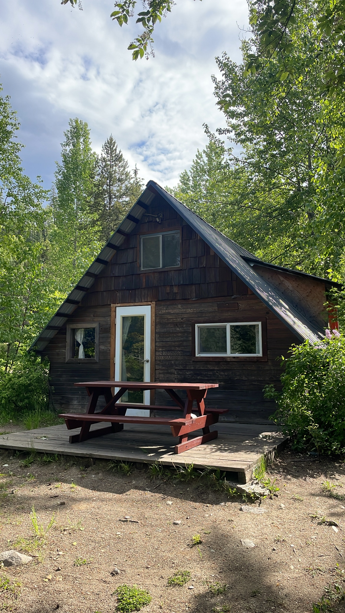 A cozy cabin is nestled among greenery, featuring an A-frame structure with a sloped roof. Large windows allow natural light to enter, and a picnic table sits on the wooden deck in front, providing an outdoor seating area.