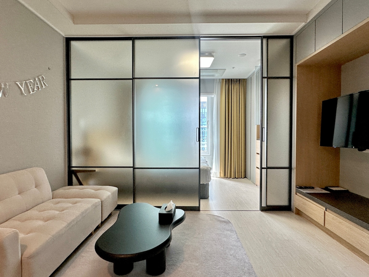 A modern living area features a light-colored sofa and a sleek coffee table. Frosted glass partitions create a separation between spaces, allowing soft natural light to filter through. A wall-mounted television is visible, while warm curtains frame the window, enhancing a cozy environment.