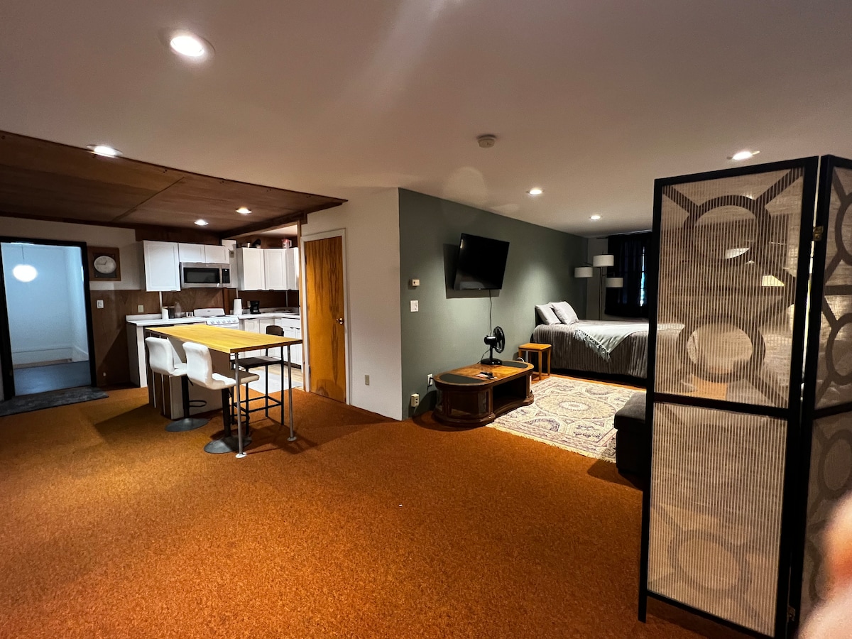 An open living space is displayed, featuring a kitchen area with seating for four. A spacious sleeping area is visible, complete with a bed and television. A decorative screen provides privacy between the living and sleeping areas, and warm lighting enhances the overall ambiance.