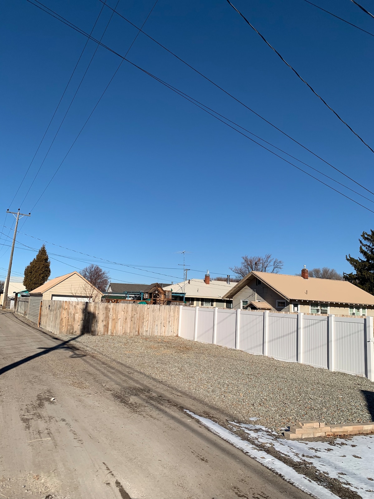 A clear blue sky is visible above a gravel area, bordered by a white vinyl fence on the right. Wooden fences are present on the left, along with several utility poles in the background. The scene captures the peaceful atmosphere of the surrounding neighborhood.