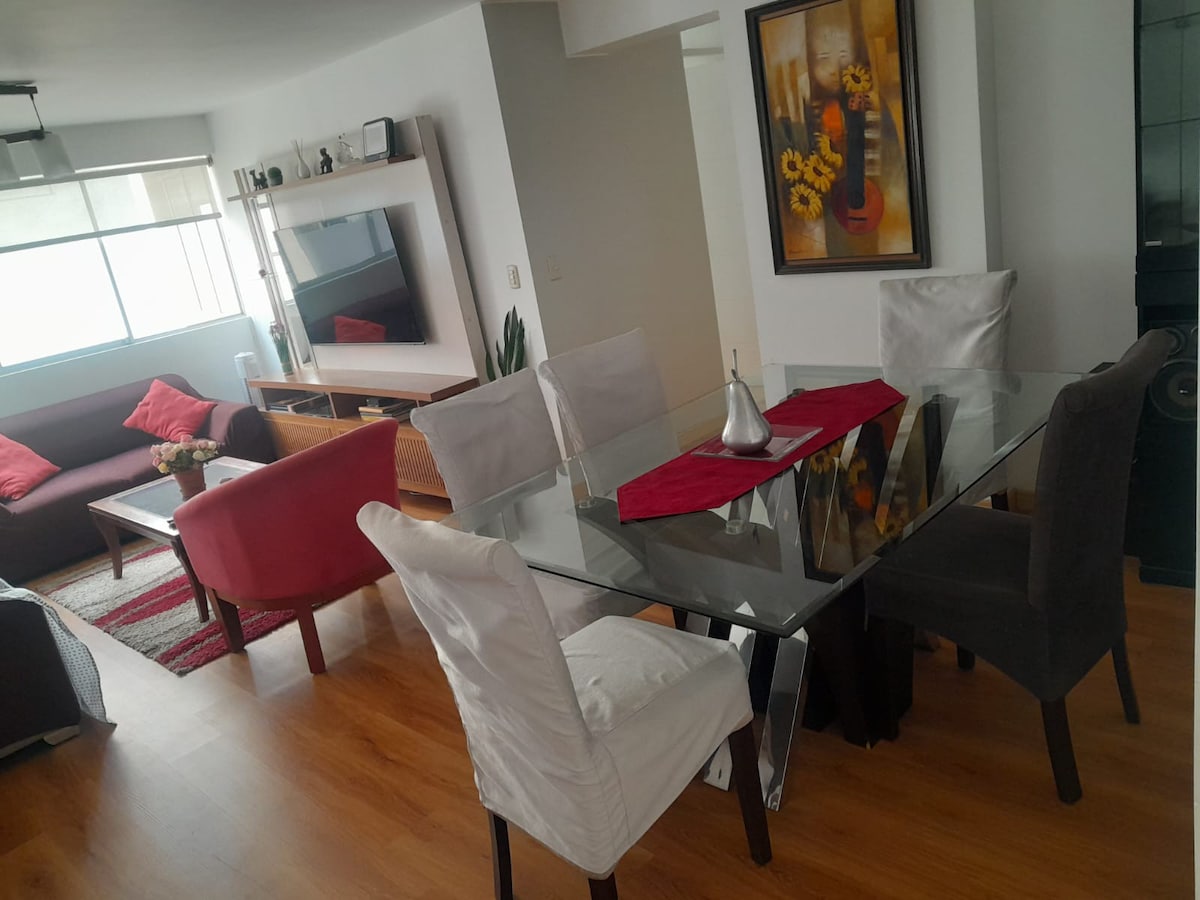 The living area features a glass dining table surrounded by six upholstered chairs. A cozy seating arrangement with a sofa and chairs in neutral tones and decorative cushions is visible. A large window allows natural light, and a piece of wall art with sunflowers adds a touch of color.