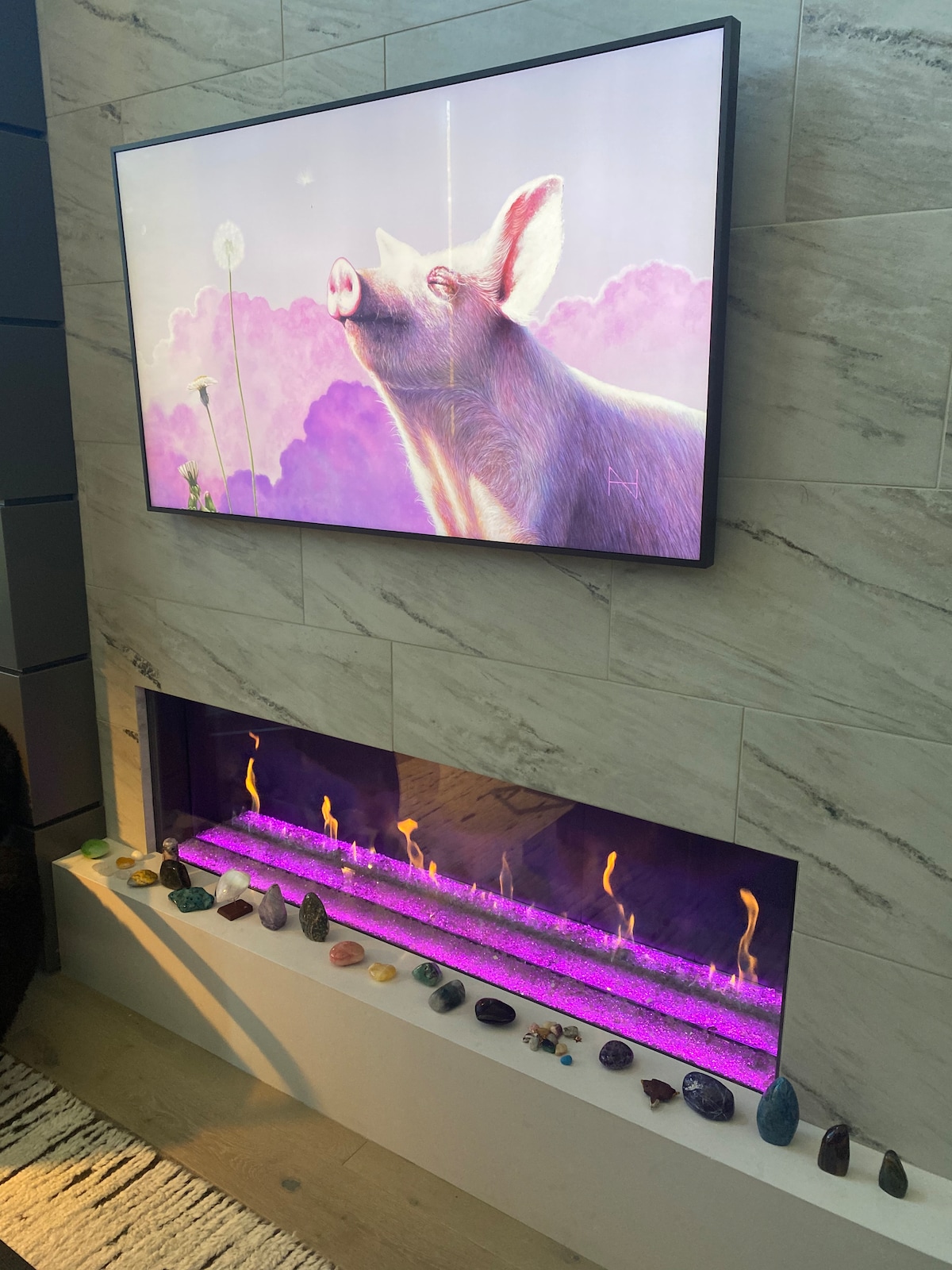A modern living area showcases a large screen displaying a whimsical image of a pig against a dreamy purple background. Below, an electric fireplace with vibrant purple lighting is lined with colorful stones, providing a cozy yet contemporary ambiance.