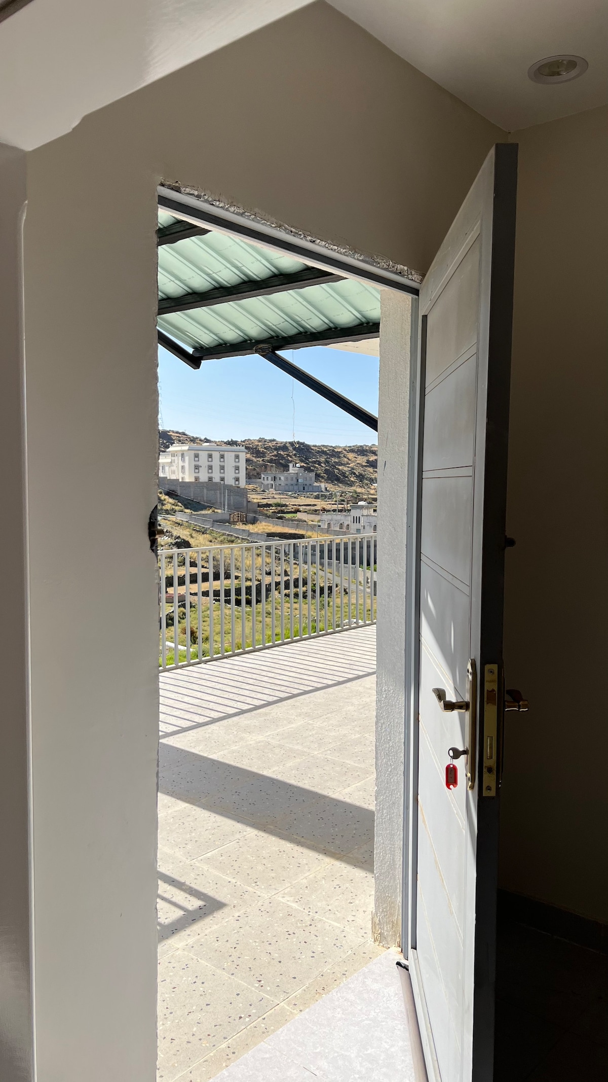 A doorway is shown ajar, leading to a balcony area with a metal railing. The landscape beyond features distant buildings and natural terrain, illuminated by daylight. The floor inside is tiled, and a portion of the ceiling is visible.