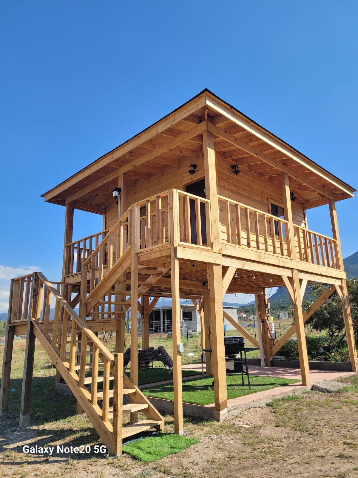 A wooden cabin is elevated on stilts, featuring a spacious deck with railings. Steps lead up to the entrance. The structure is surrounded by open space and mountains are visible in the background under a clear blue sky.