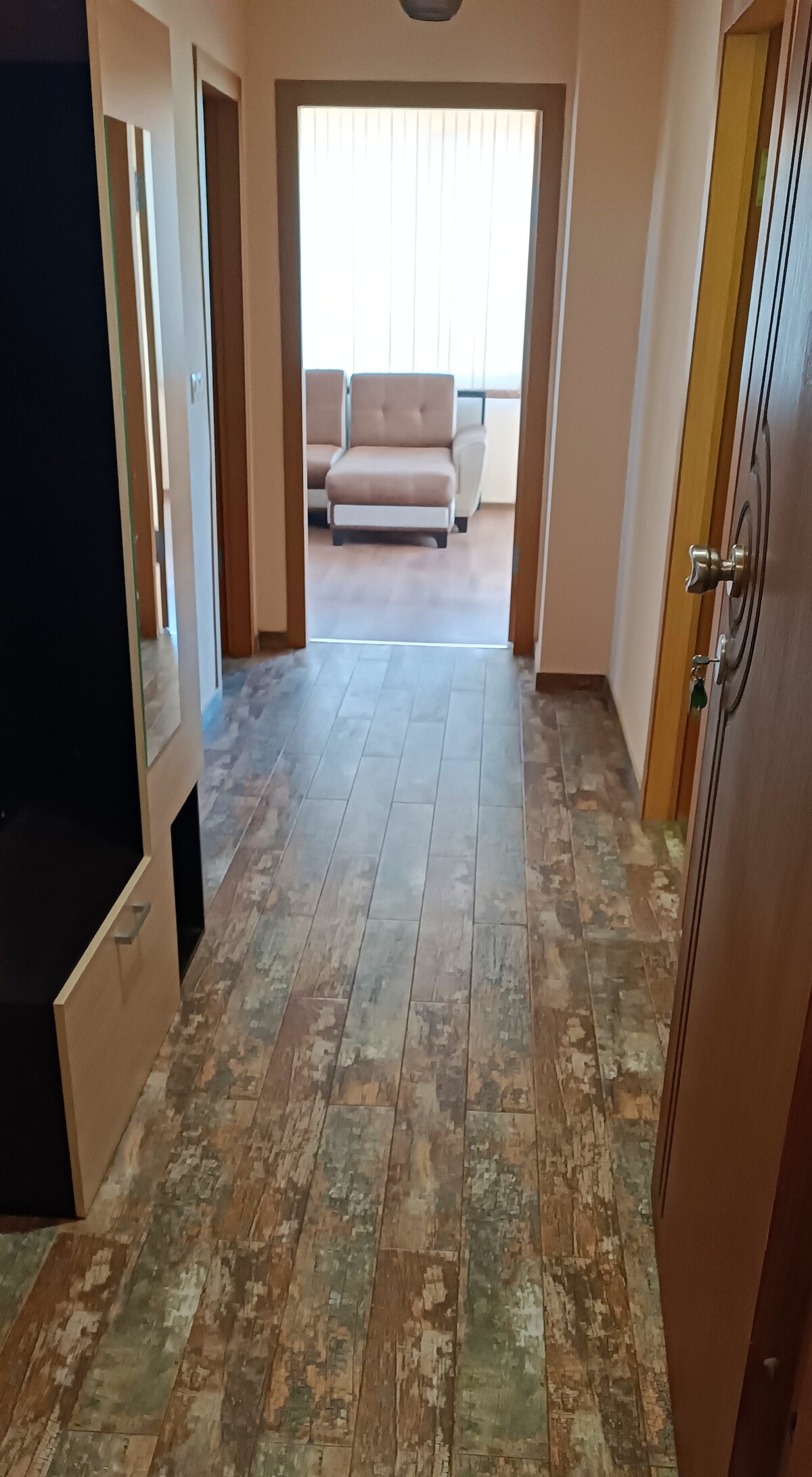 A hallway is presented, featuring a tiled floor designed to resemble wood. Doorways lead to rooms on either side, with a window at the end allowing natural light. A pair of light-colored seating arrangements is visible in the adjacent room.