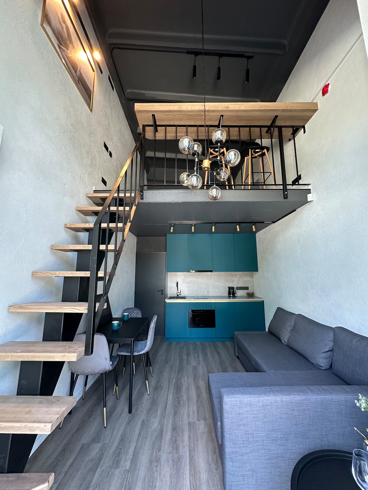 A modern loft space features a striking open layout with high ceilings and a wooden mezzanine. A sleek kitchen with teal cabinetry is visible, complemented by a dining area set for four. A comfortable gray sofa is positioned near the staircase, enhancing the stylish ambiance.