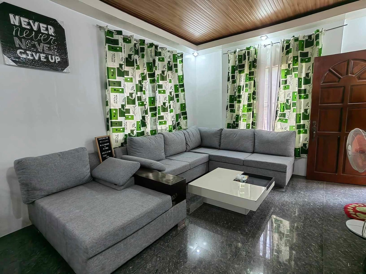 A spacious living area features a large sectional sofa positioned around a coffee table. Vertical curtains with a green and white geometric pattern frame the window, while natural light brightens the polished floor. A decorative wall sign adds a motivational touch.