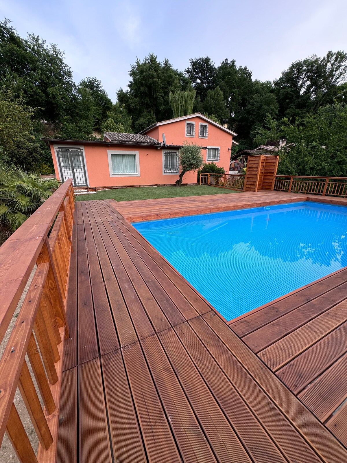 A wooden deck surrounds a sparkling blue pool, providing a relaxed outdoor space. A charming pink house is positioned in the background, flanked by greenery. A path leads to the house, visible behind the pool, creating a harmonious connection between the home and nature.