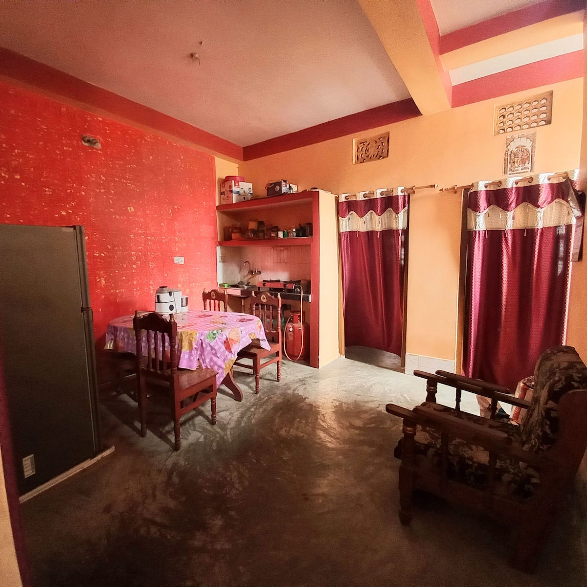 A cozy living area is shown, featuring a small dining table surrounded by four chairs and a kitchenette equipped with shelves for storage. Two framed windows with rich maroon curtains complement the warm orange walls, while a comfortable armchair adds to the inviting space.