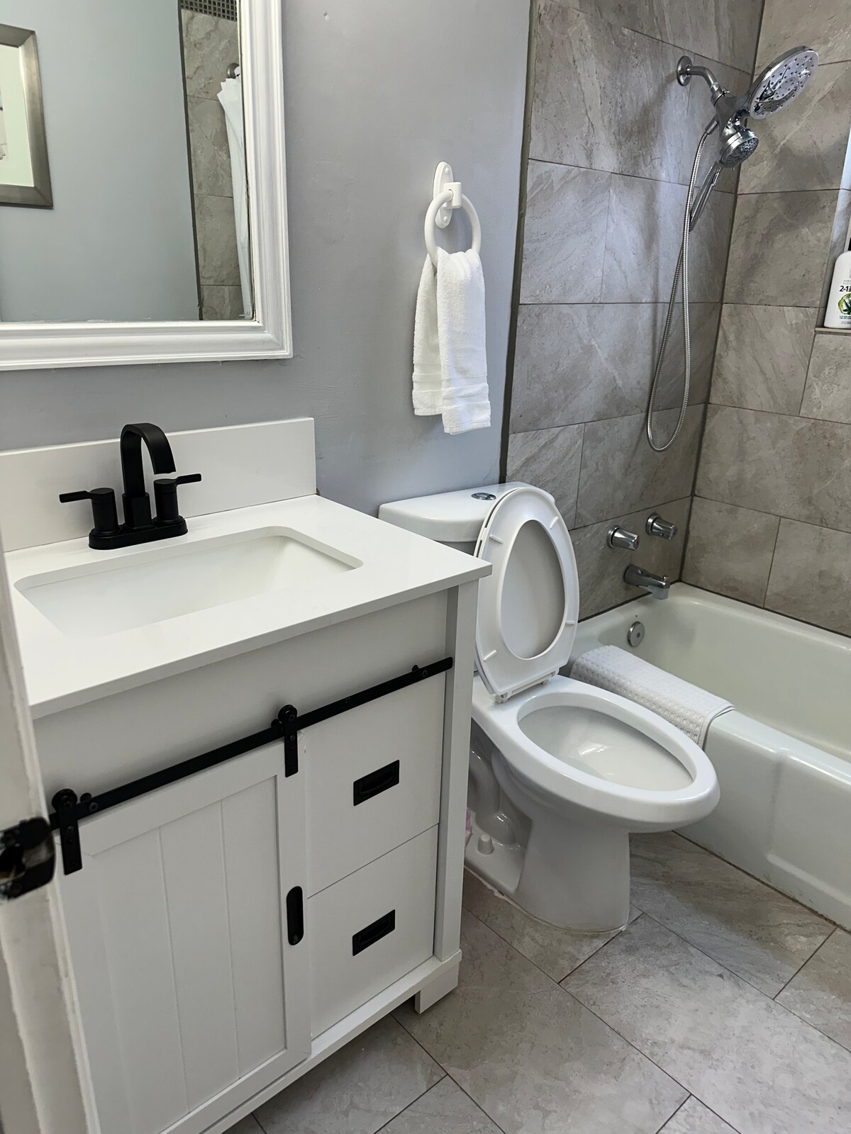 A modest bathroom features a white vanity with a sleek sink and stylish faucet. The neutral-toned tiled walls encompass a bathtub with a showerhead above. Clean towels hang on a rack, and a mirror reflects natural light in the space.