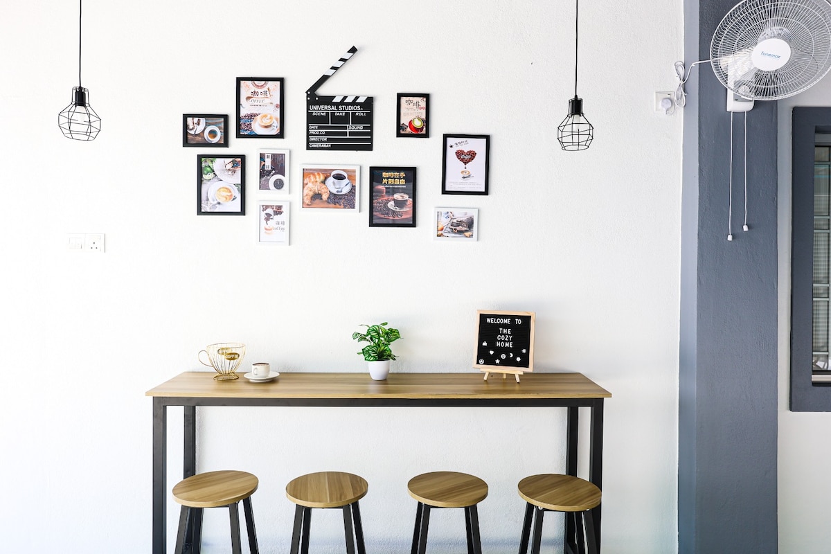 A narrow wooden table is positioned against a bright wall, accompanied by four stools. Above the table, a collection of framed photographs and decorative items are arranged. Two pendant lights hang from the ceiling, while a small plant adds a touch of greenery to the setting.
