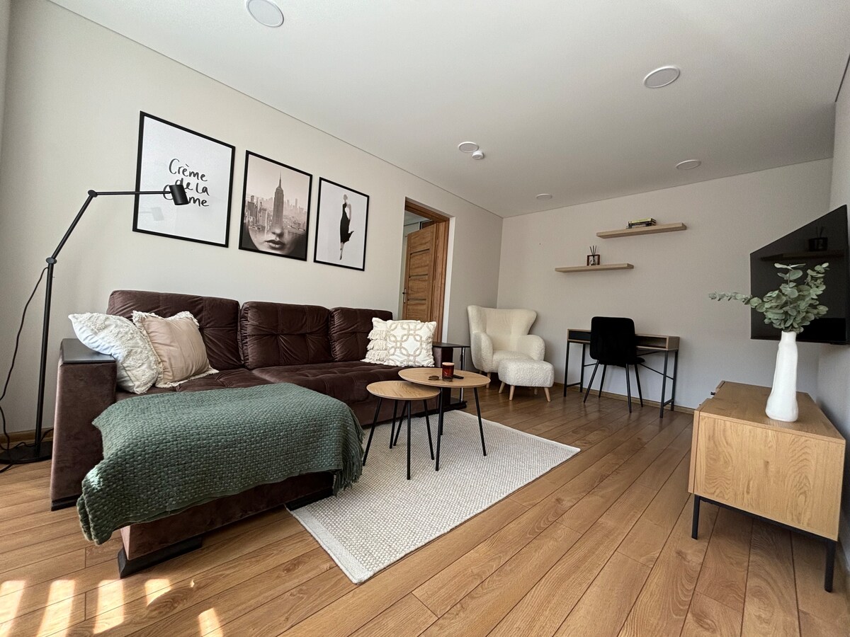 The living area features a dark sectional sofa adorned with decorative pillows and a soft throw. A round coffee table rests on a light area rug, complemented by a modern armchair and a desk against the wall. Framed art adds to the minimalist decor.