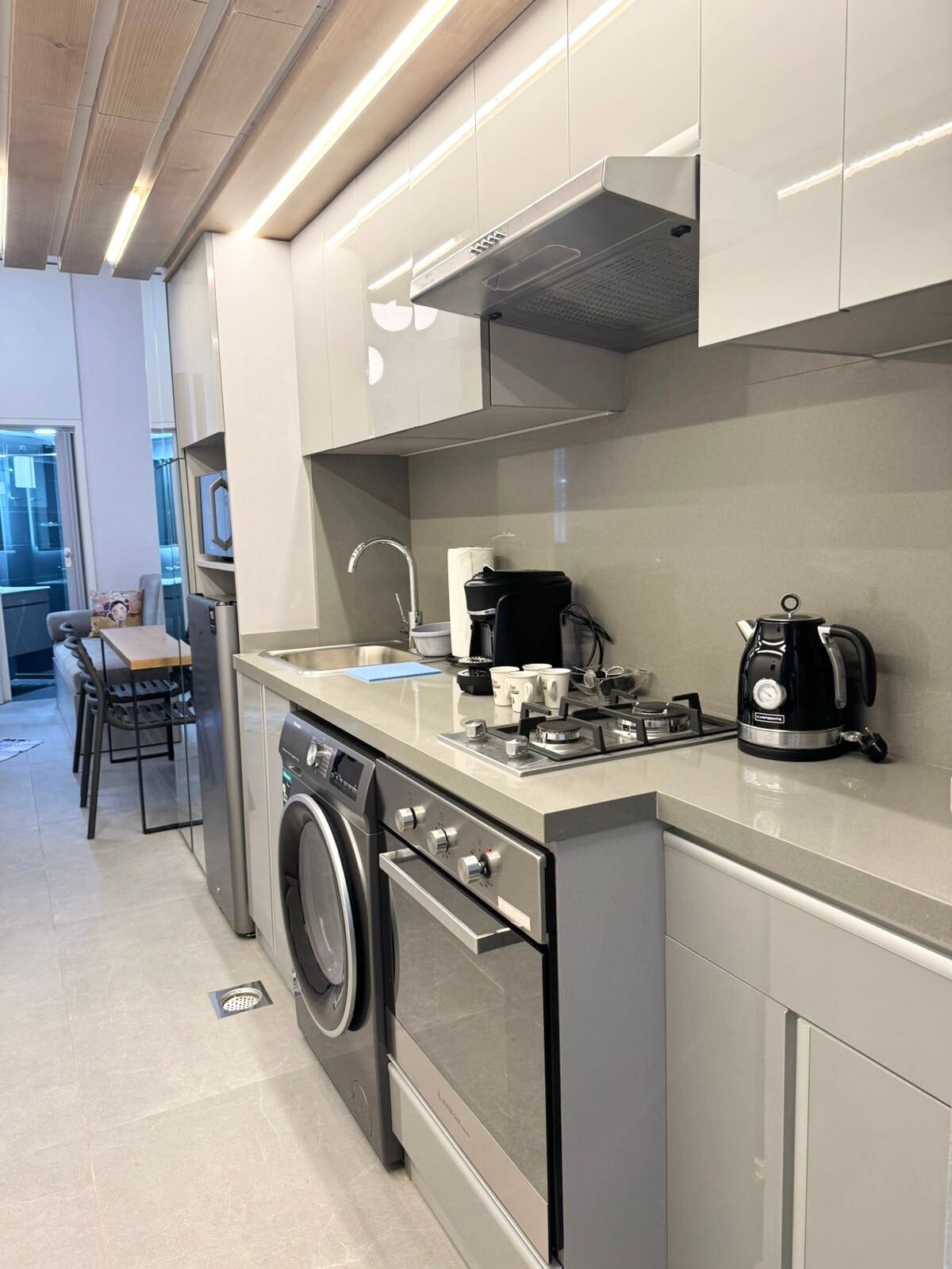 A sleek kitchenette is showcased with modern cabinetry and a countertop. Featured appliances include a washing machine, oven, microwave, espresso machine, and kettle. Cutlery and dishware are neatly arranged, providing a functional space for meal preparation. Natural light enters from a nearby window.