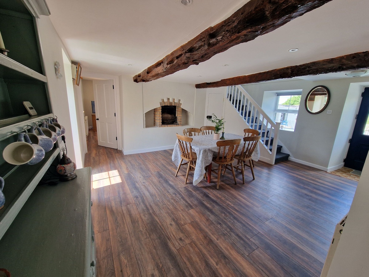 The dining area features a round table surrounded by six wooden chairs, positioned under exposed oak beams. Light from a nearby window enhances the warm wood tones of the floor. A fireplace is visible in the background, adding character to the space.
