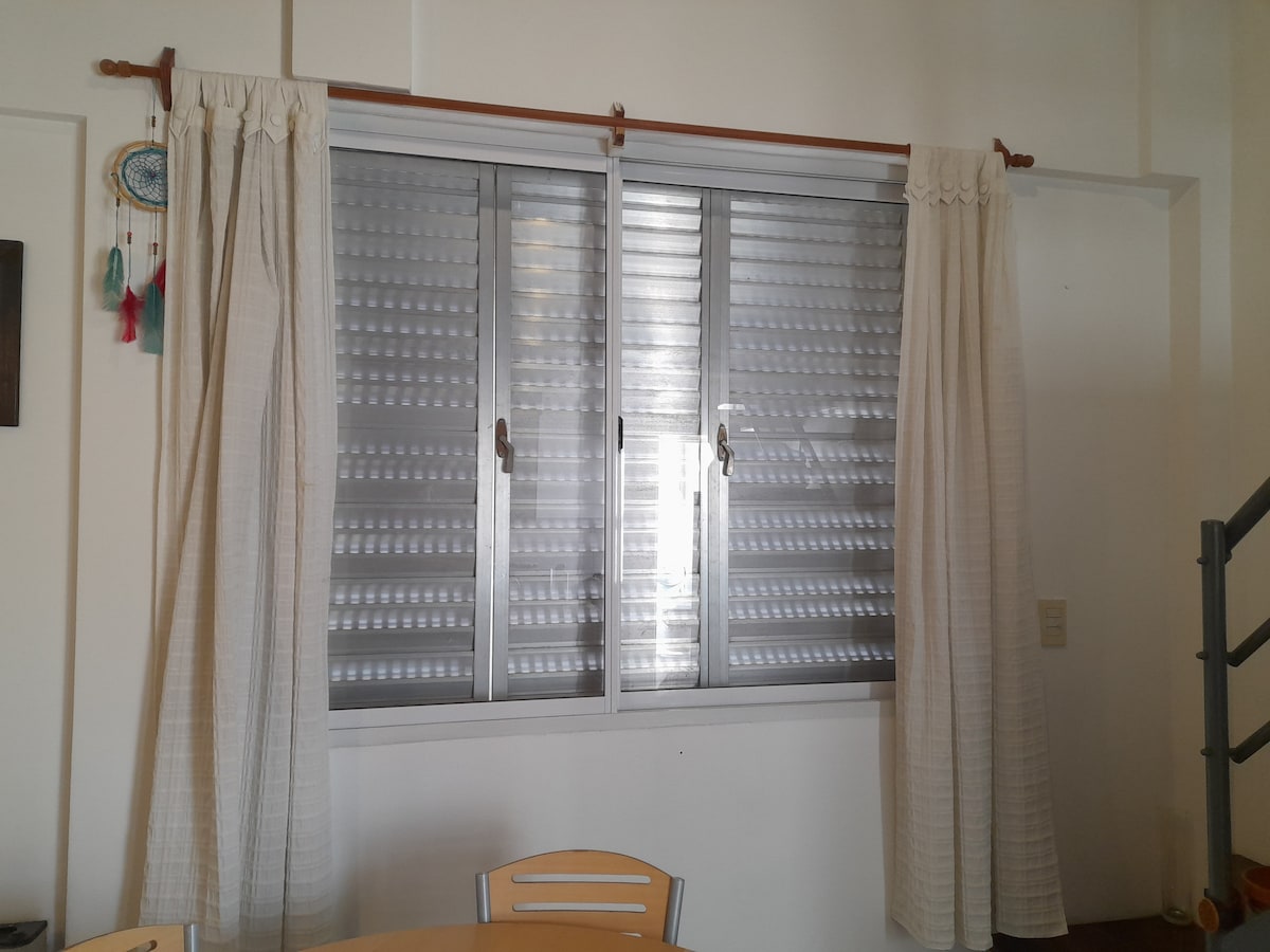 A window with multiple sliding panels is framed by light-colored, sheer curtains. Natural light is allowed to filter through, illuminating the nearby dining area with a round table and simple chairs.