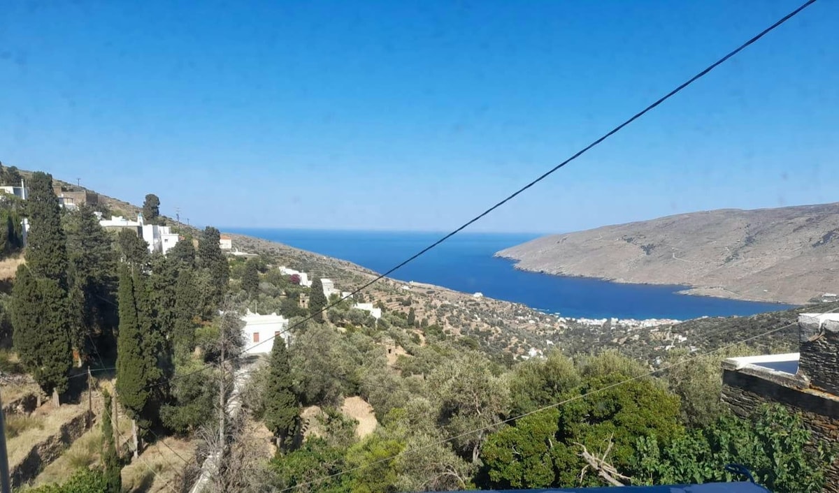 A panoramic view of the coastline is depicted, showcasing a blend of lush greenery and rocky terrain leading down to a sparkling blue sea. The clear sky frames the scene, while distant hills and white structures dot the landscape, creating a serene coastal atmosphere.