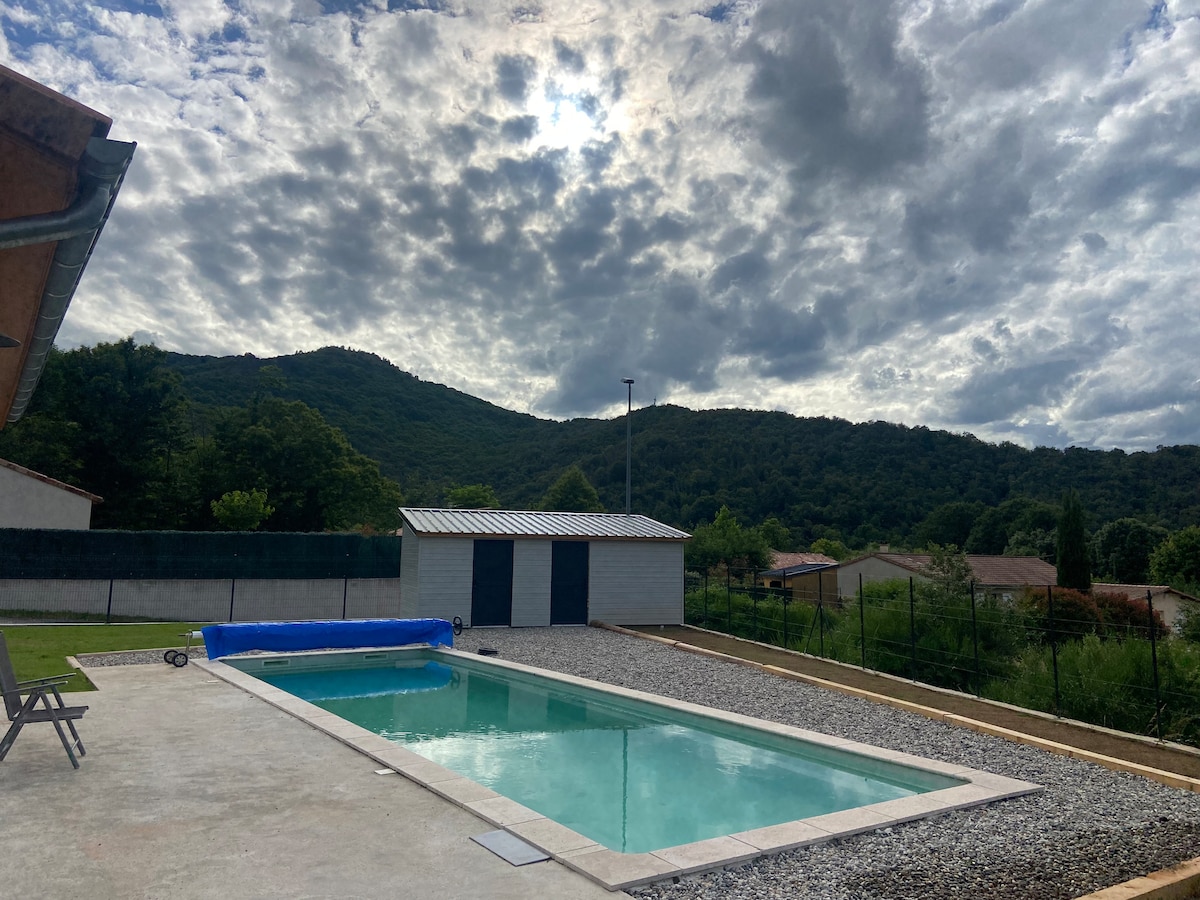 A private saltwater pool is set against a backdrop of lush mountains, with a blue cover partially rolled beside it. Gray stone surrounds the pool area, and a spacious deck offers seating options. The sky is filled with dramatic clouds, creating a peaceful atmosphere.