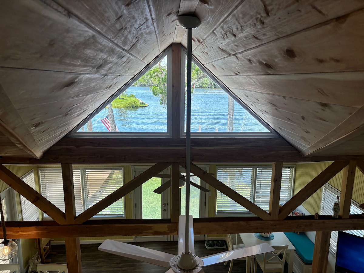 The image captures a spacious interior view from the loft, showcasing exposed wooden beams and a ceiling fan. Large windows provide an unobstructed view of the tranquil lagoon, with greenery visible outside. Natural light illuminates the area, enhancing the inviting atmosphere.