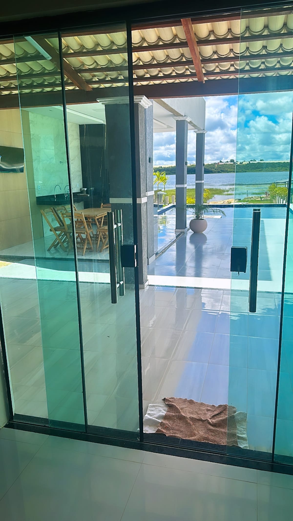 A large glass door offers a view of the outdoor dining area next to a serene body of water. Natural light enters the space, reflecting off modern tiled floors. A soft rug is positioned at the entrance, contributing to the welcoming atmosphere.