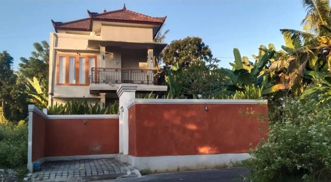 The villa is showcased from the street, displaying a two-story design with a distinctive roof. Lush greenery surrounds the exterior, while the orange wall defines the property. Large glass windows provide glimpses of the interior, emphasizing the connection to the natural setting.