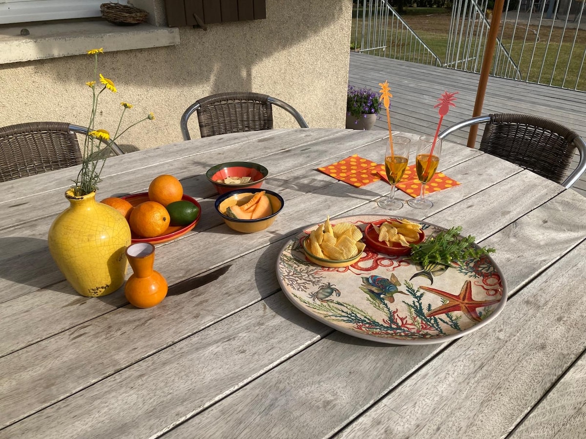 A wooden table is set outdoors, displaying a vibrant collection of fruits and snacks. A decorative plate features assorted chips, while colorful bowls hold slices of melon. Two glasses await refreshments, and a yellow vase filled with fresh flowers adds a touch of color.