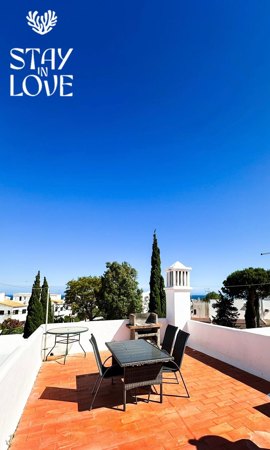 A spacious terrace is showcased, featuring a dining table and chairs arranged neatly on terracotta tiles. Lush trees and a distant view of the ocean are visible against a clear blue sky. A barbecue area is included, enhancing the outdoor space.