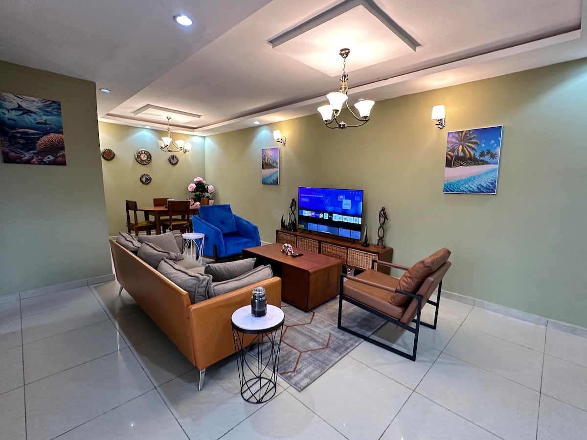A welcoming living area is designed for comfort, featuring a brown leather sofa, contrasting armchair, and a coffee table atop a soft area rug. A large TV is mounted on the wall, illuminated by ceiling lights that enhance the space's ambiance.