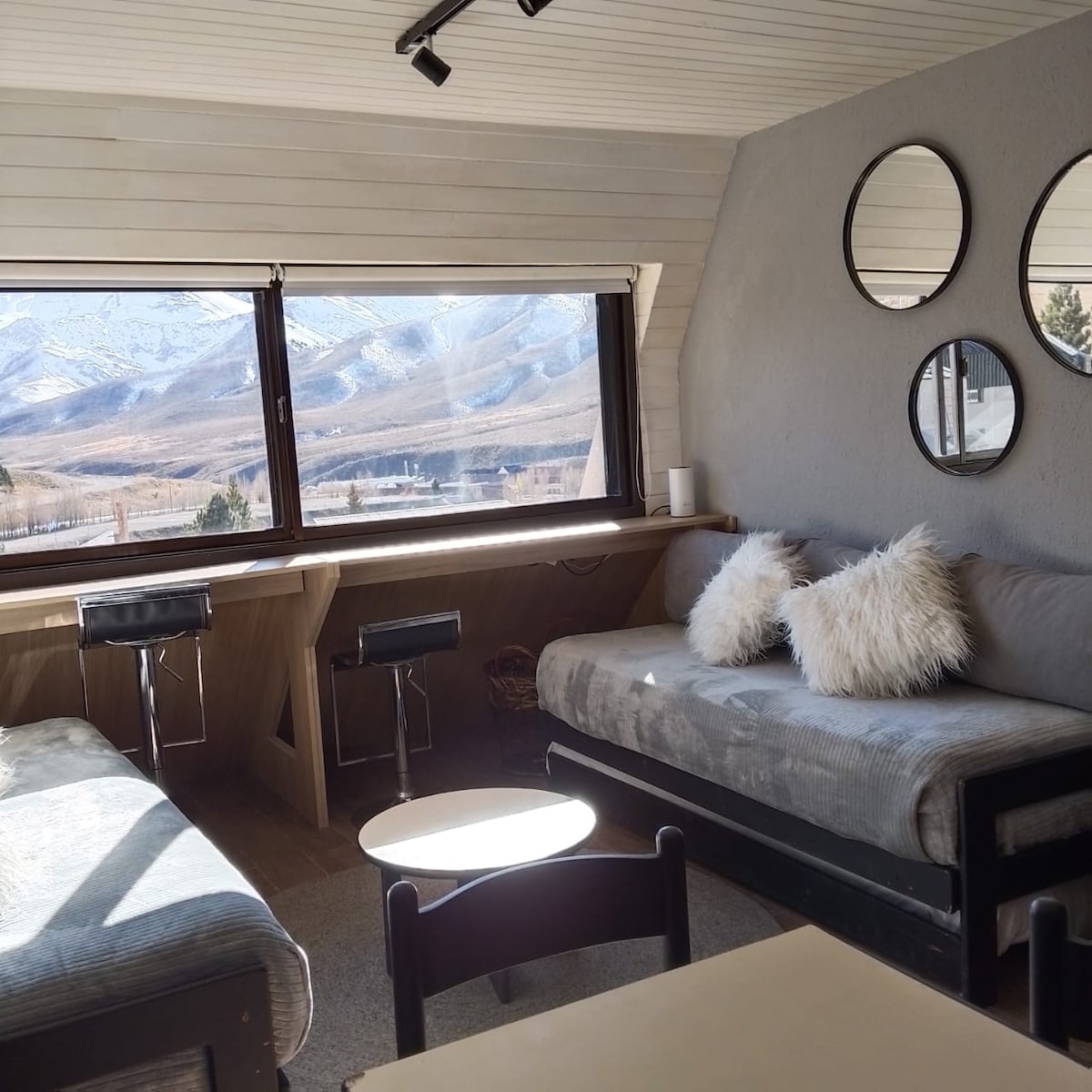 A modern living area features large windows showcasing mountain views. Light-colored walls create an open feeling, while two plush sofas and a round table provide inviting seating options. Decorative mirrors adorn the wall, reflecting natural light throughout the space.