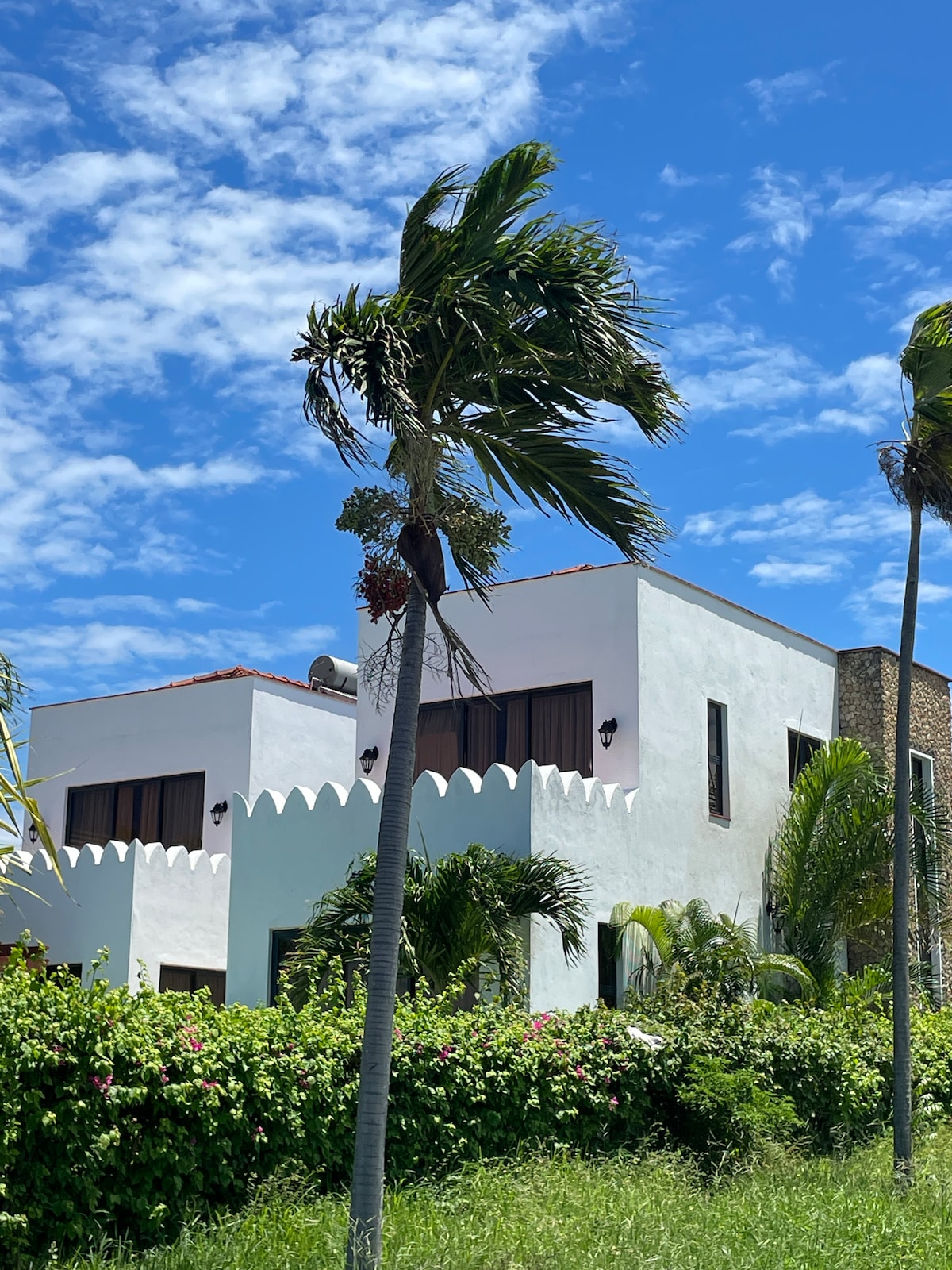 The exterior of the holiday home is showcased with a backdrop of a bright blue sky and scattered clouds. Lush greenery and vibrant plants surround the property, while decorative features highlight the unique architecture. Palm trees gently sway in the breeze, creating a tropical ambiance.