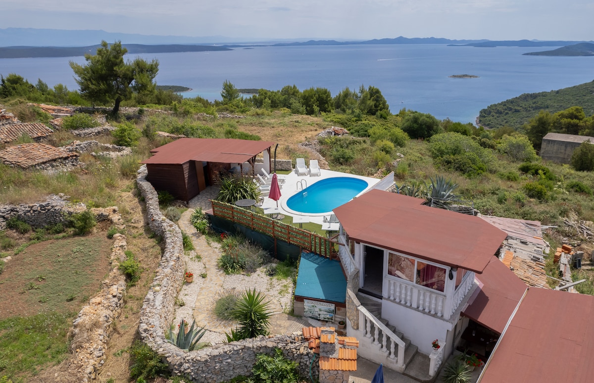 An elevated view captures a serene landscape featuring a private swimming pool surrounded by lounge chairs. Rustic buildings with terracotta roofs blend harmoniously with the lush green surroundings, offering a glimpse of the coastline and distant islands under a clear sky.