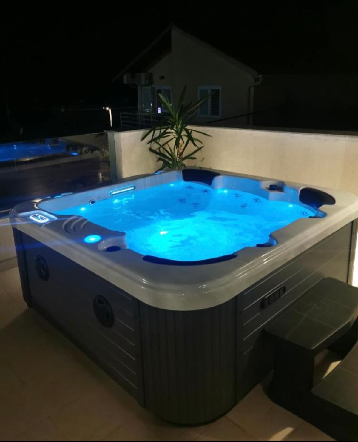 A hot tub is featured on an outdoor terrace, illuminated with soft blue lighting. The space is surrounded by a glass railing, providing an open view. A small seating area is nearby, enhancing the relaxing ambiance.