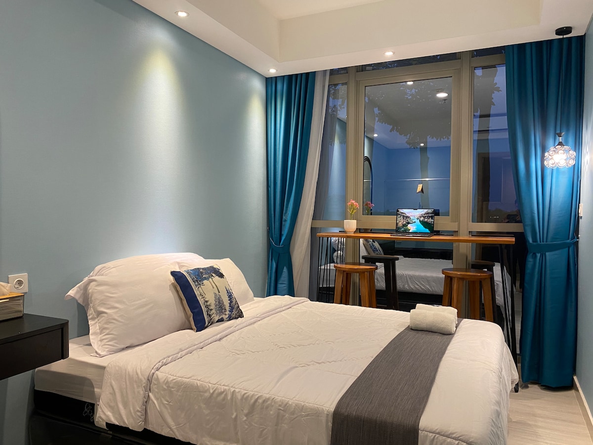 A well-lit bedroom features a spacious bed with white linens and a decorative blue throw pillow. Two high chairs are positioned at a small table near the large window, framed by blue curtains. Soft lighting enhances the calming atmosphere of the room.
