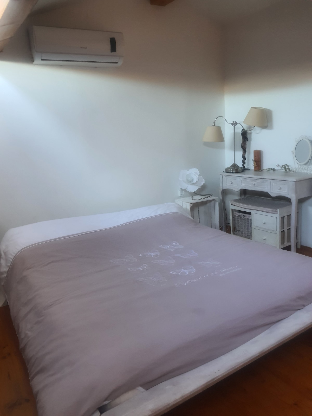 A simple yet comfortable sleeping area features a spacious bed with a soft lavender coverlet. Natural light filters in, illuminating a small vanity area with a mirror and decorative accents. A gentle ambiance is created by the lamps perched on either side.