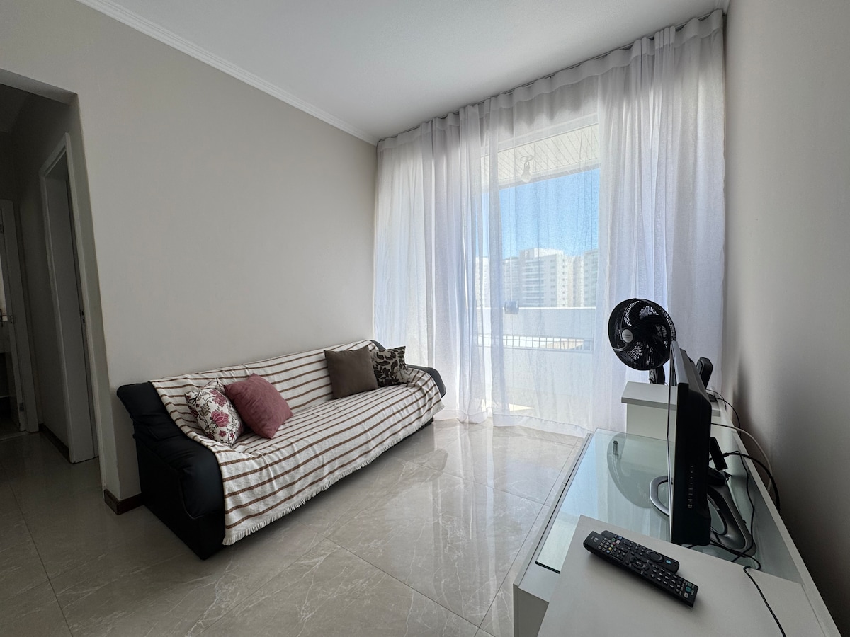 A bright living area features a striped sofa with colorful cushions, positioned beside a large window that allows natural light to fill the space. A sleek desk and fan are also visible, contributing to a functional and comfortable atmosphere.