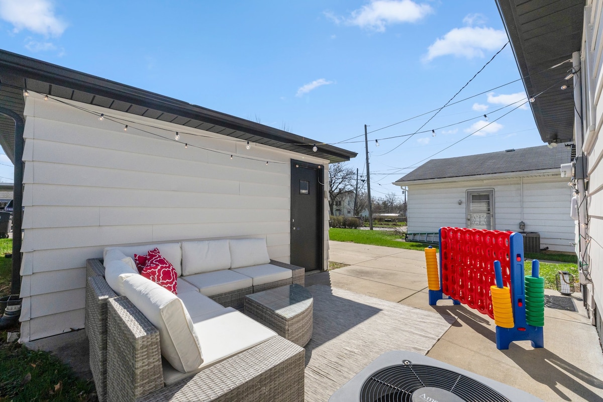 A private patio is featured, showcasing a spacious outdoor seating area with a light-colored sectional and decorative pillows. Nearby, a multi-colored oversized game set stands ready for entertainment. Clear skies and overhead string lights enhance the inviting space.