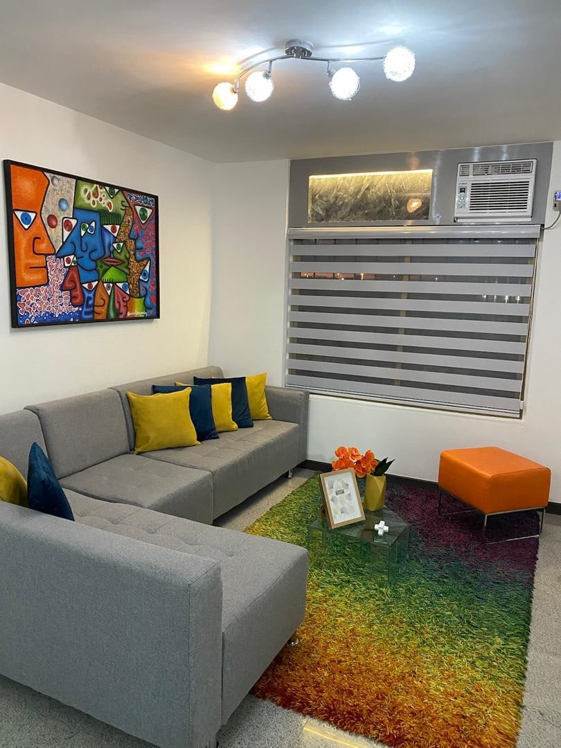 A contemporary living area features a gray sectional sofa adorned with colorful cushions. A vibrant artwork hangs above, and a small orange accent chair complements the space. A modern rug in gradient colors covers the floor, with a glass coffee table displaying a framed photo and flowers.