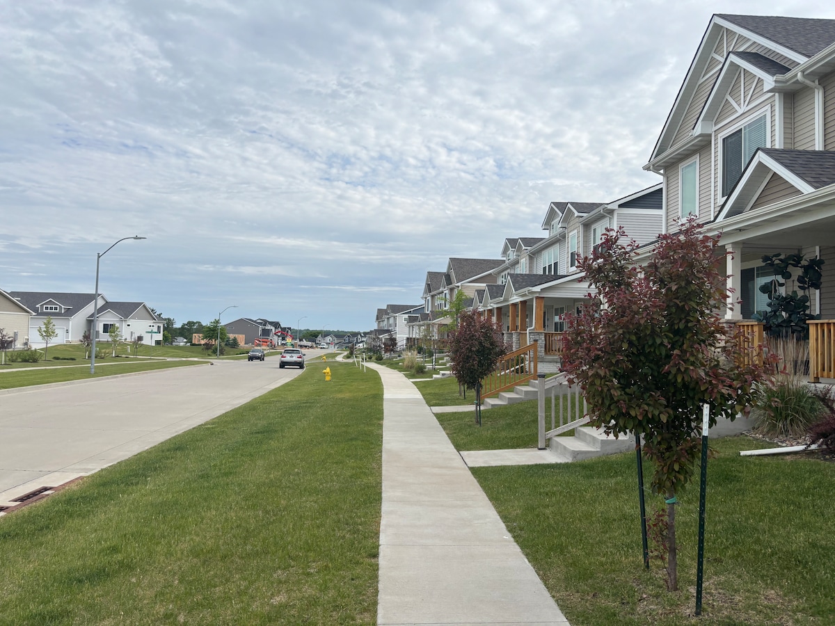 A suburban street is captured, lined with well-kept homes featuring porches and manicured lawns. A wide sidewalk runs parallel to the street, which is clear of traffic. Trees with green foliage are positioned along the sidewalk, offering a sense of greenery to the neighborhood.