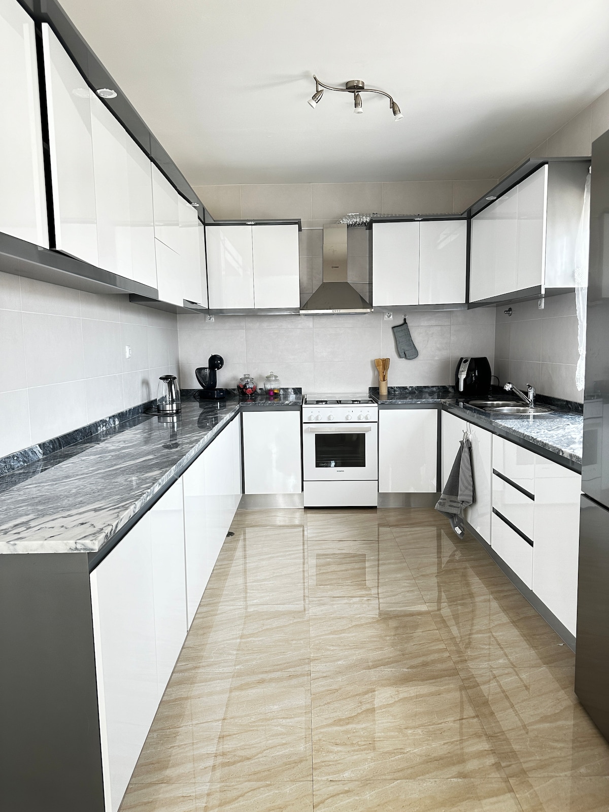 A modern kitchen is displayed with sleek white cabinets and a polished marble countertop. Stainless steel appliances, including a gas stove and a coffee maker, are present. The tile flooring reflects light, enhancing the spacious feel of the kitchen.