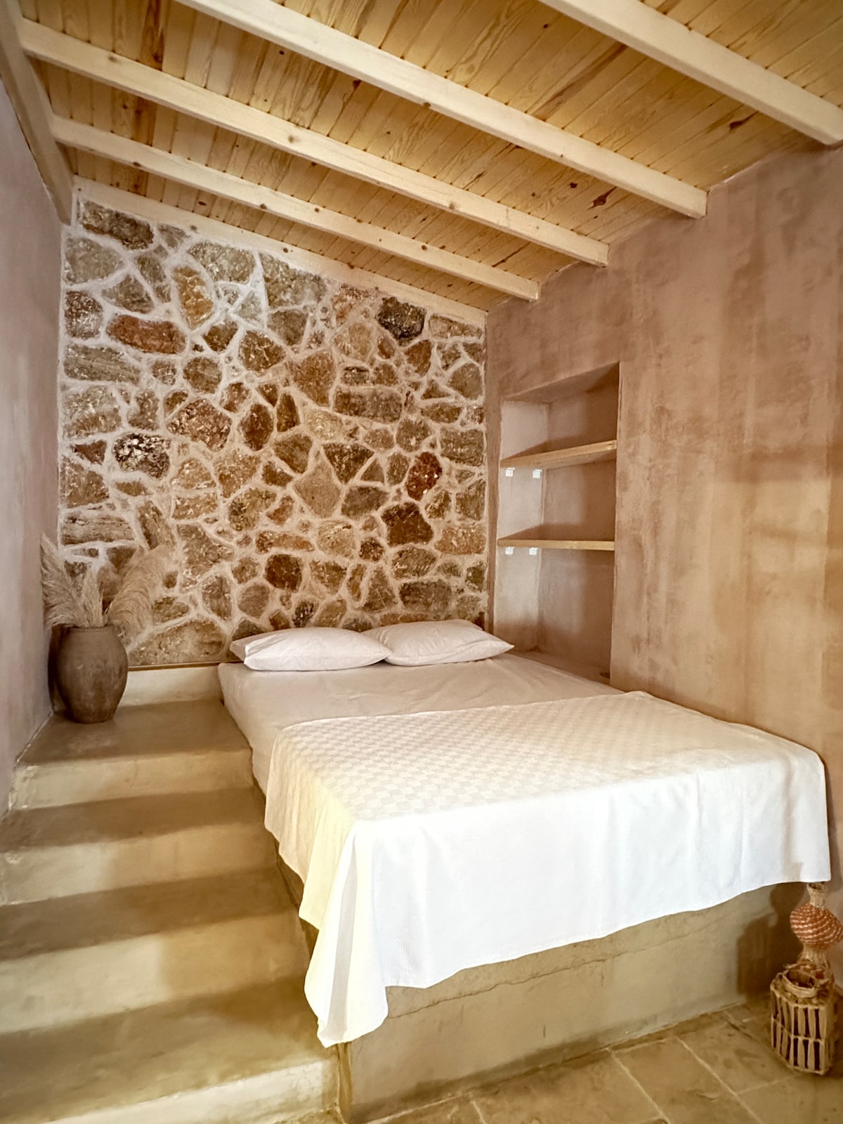 A sleeping area features a spacious bed with a white bedspread, positioned against a stone accent wall. Natural light enters the space, highlighting the wooden ceiling and minimalist shelves on one side. Decorative elements include a vase with dried plants, enhancing the serene environment.