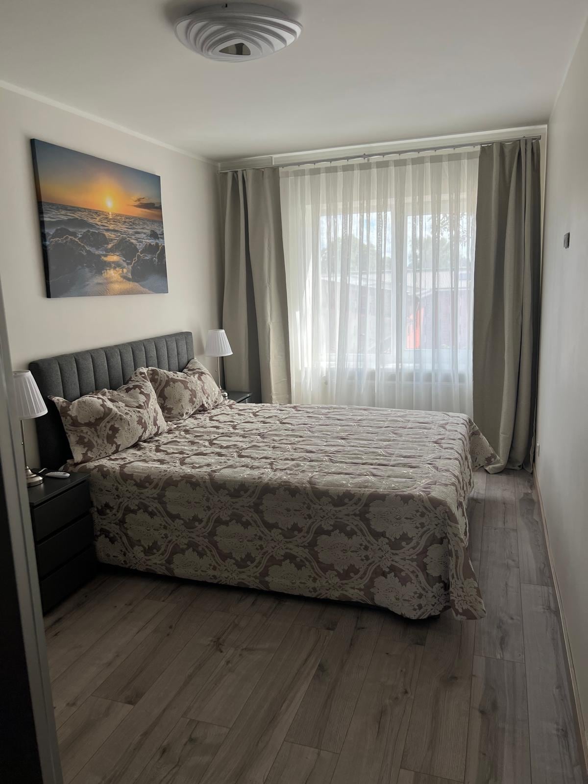 A comfortable bedroom features a spacious bed adorned with a patterned blanket. Soft natural light filters through sheer curtains, illuminating the room. Two bedside lamps provide additional lighting, and a landscape artwork is displayed above the bed, enhancing the serene ambiance.