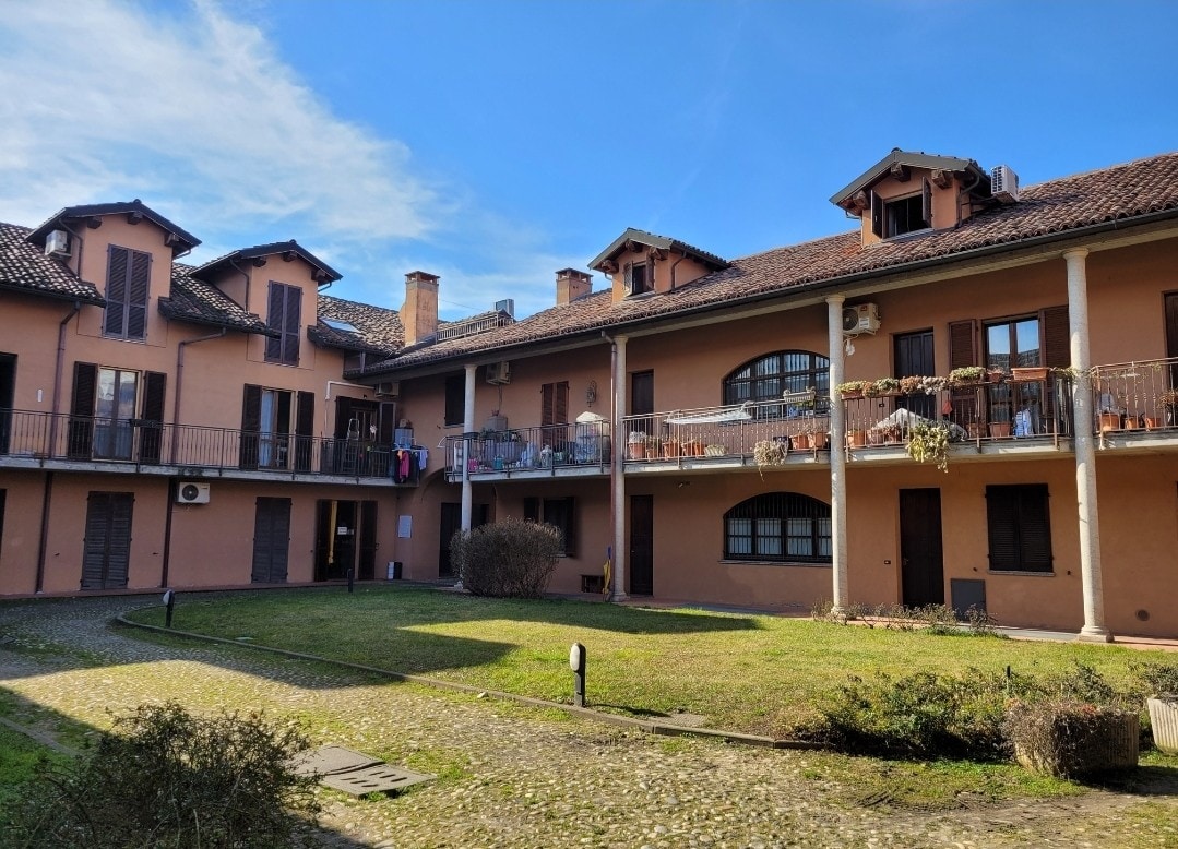 The building features a warm exterior, showcasing multiple levels with gabled roofs and balconies adorned with potted plants. A grassy courtyard is framed by walkways, and the sunlight casts a soft glow on the serene scene. Clothes are hung on lines, suggesting a home-like atmosphere.