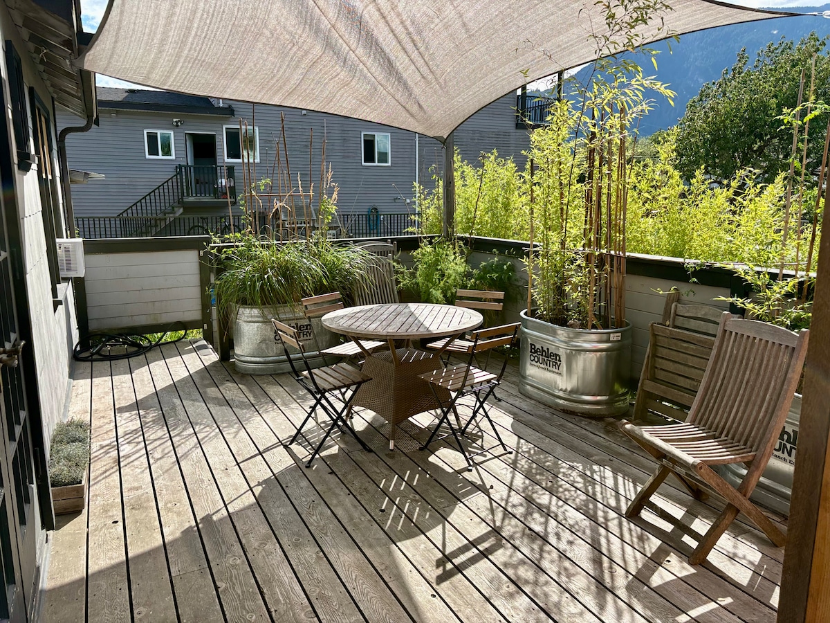 A spacious deck is covered by a shade sail, accommodating a round table surrounded by folding chairs and wooden loungers. Potted bamboo plants and greenery enhance the natural ambiance, while the backdrop features distant mountains and neighboring buildings.