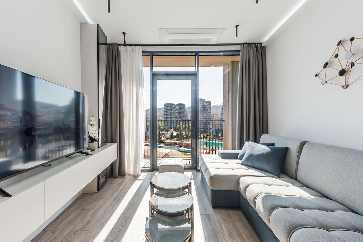 A modern living area showcases a comfortable sectional sofa and a sleek television mounted on the wall. Large floor-to-ceiling windows allow natural light to fill the space, offering a view of the outdoor landscape and nearby buildings.