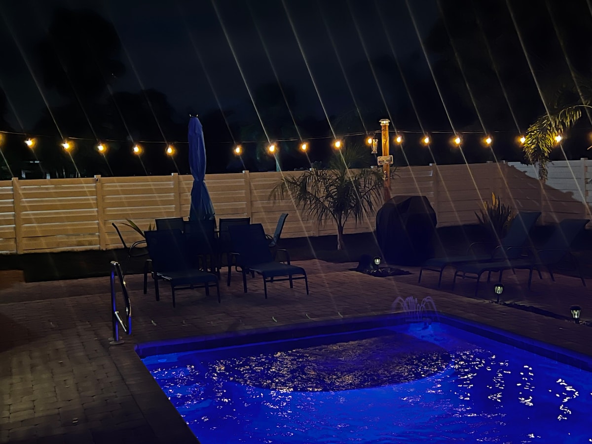 A tranquil pool area is highlighted by soft blue lighting, complemented by string lights overhead. Several lounge chairs surround the pool, while tall umbrellas provide shade. The backdrop features a wooden fence and lush greenery, enhancing the serene evening ambiance.